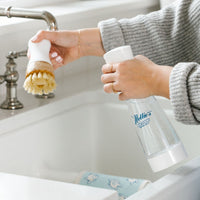 Person using Nellie's Ninety-Nine to remove bacteria on their Nellie's Kitchen Brush over a kitchen sink with a Nellie's Swedish Dishcloth in the background