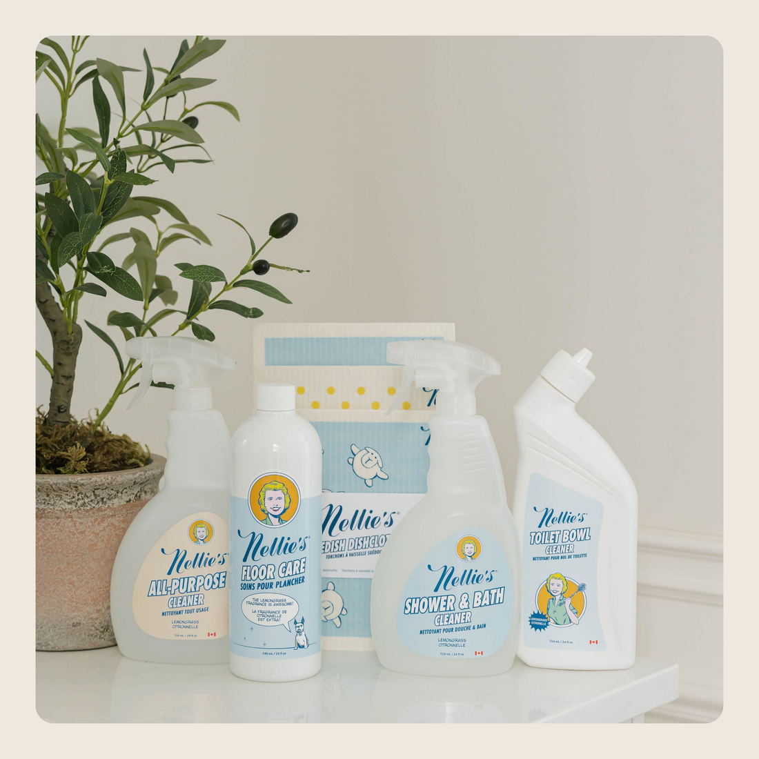 A set of Nellie's cleaning products arranged on a white shelf next to a potted plant. The products include a floor cleaner, a hand wash bottle, a shower and bath cleaner, and a water & stain cleaner, with a branded box behind them.