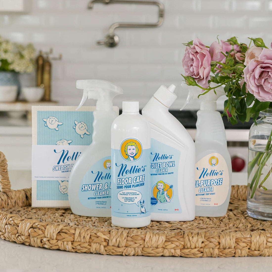 A set of Nellie's All Purpose Cleaning products arranged on a woven tray in a bright kitchen setting, with pink roses in a vase nearby.