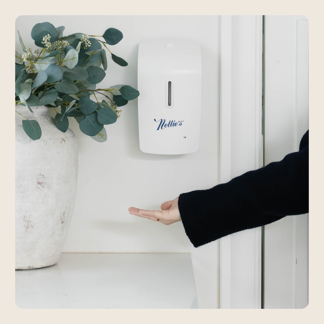 A white wall-mounted hand sanitizer dispenser with the brand name 'Hollie's' in blue script, shown next to a person's outstretched hand in a clean indoor setting.