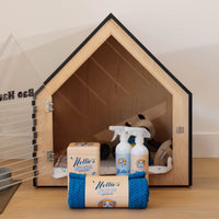 A wooden house-shaped dog crate with a clear door and black trim, shown on a tabletop with pet accessories in front. Visible items include a rolled blue rope toy with a Nottie's label, two white spray bottles, and a box.