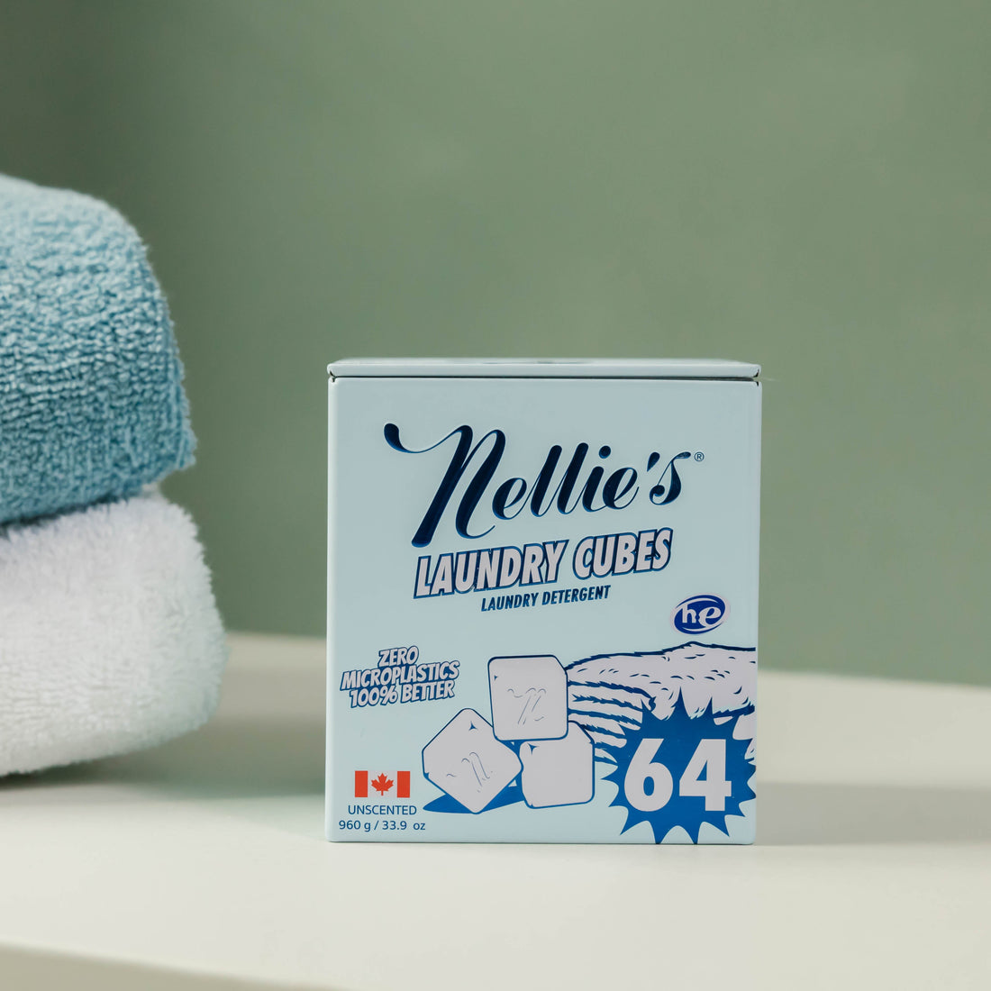 A box of Nellie's Laundry Cues liquid detergent placed on a white surface next to stacked blue and white towels, with a soft green wall in the background.