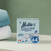A box of Nellie's Laundry Cues liquid detergent placed on a white surface next to stacked blue and white towels, with a soft green wall in the background.