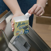 A person in a white top and blue jeans holds a box of Nellie's Dish Cubes in their hands, placing a white cube into a stainless steel dishwasher dispenser.
