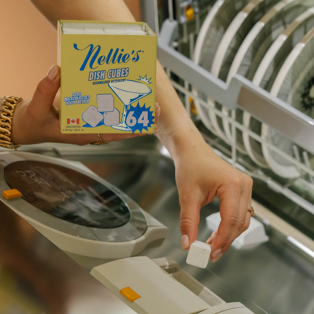 A person holds a box of Nellie's Dish Cubes and drops a single cube into a dishwasher, with dishes visible in the rack in the background.